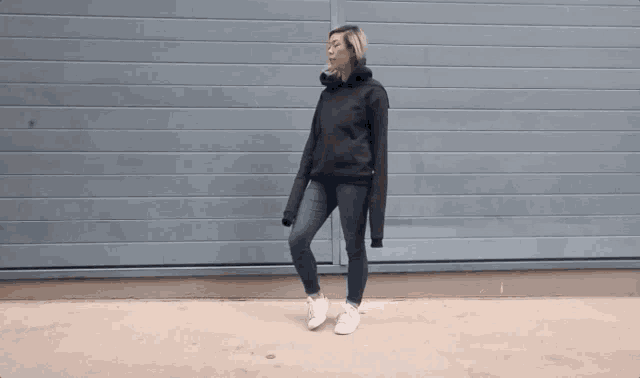 a woman in a black hoodie is standing in front of a gray wall