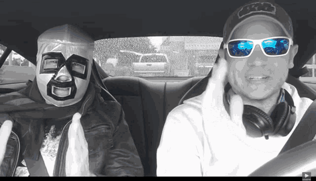 two men wearing masks and sunglasses are sitting in a car and one has a hat that says fox
