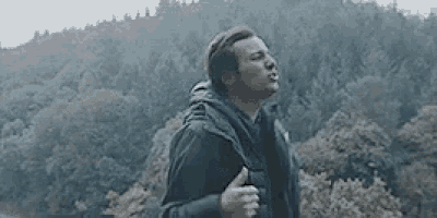 a man is standing in front of a forest holding his chest .