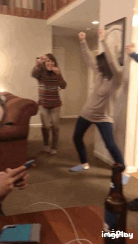 two women are dancing in a living room while someone holds a phone