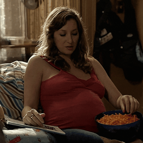 a pregnant woman in a red tank top sits on a couch eating macaroni and cheese
