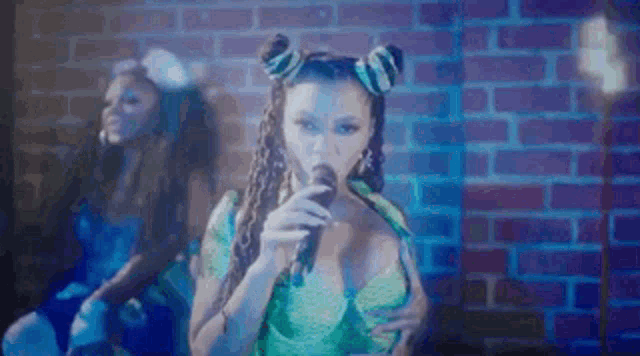a woman is singing into a microphone in front of a brick wall while another woman looks on .