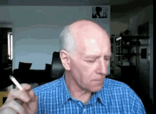 a bald man in a blue plaid shirt is smoking a cigarette in a living room .