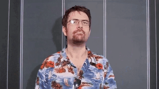 a man with glasses and a hawaiian shirt is standing in front of a wall .