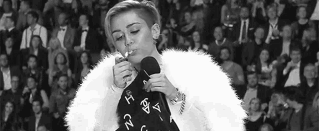 miley cyrus is smoking a cigarette in front of a crowd while wearing a fur coat .