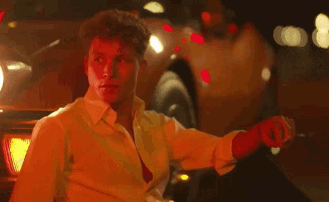 a man in a yellow shirt is sitting in front of a car at night smoking a cigarette .