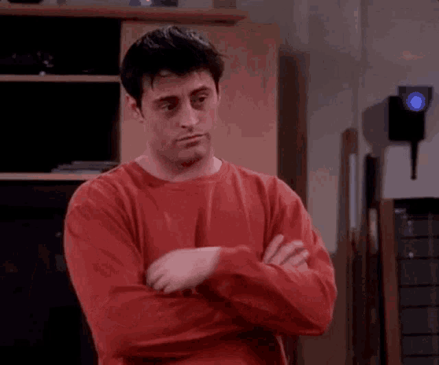 a man in a red shirt is standing in a living room with his arms crossed and looking at the camera .