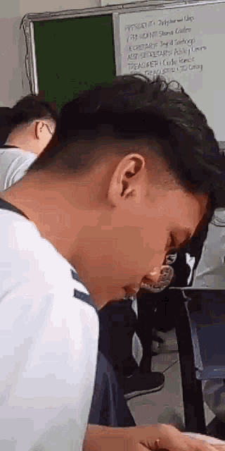 a young man is sitting in a classroom with his eyes closed and a whiteboard in the background .