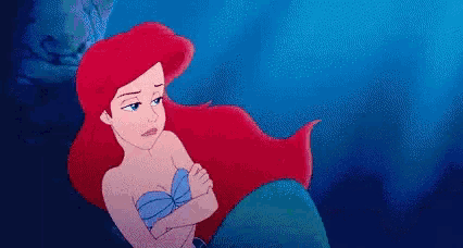 ariel from the little mermaid is sitting on the shore of the ocean with her arms crossed .
