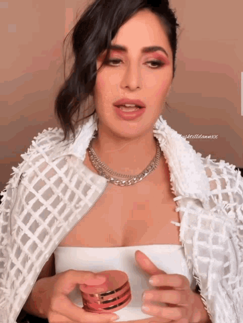 a woman wearing a white jacket and a necklace holds a bottle