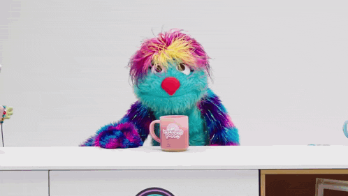 a colorful stuffed animal is sitting at a table with a pink mug that says ' sunny day ' on it