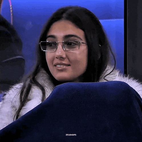 a woman wearing glasses and a fur coat is sitting on a blue chair