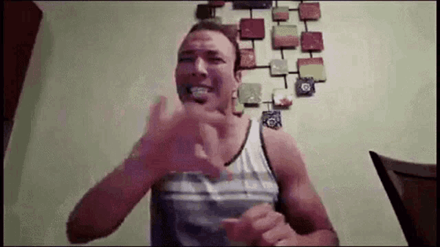 a man in a tank top is making a funny face while sitting in front of a wall with a painting .