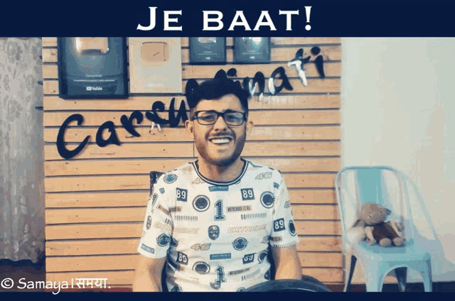 a man wearing glasses stands in front of a wooden wall that says je baat