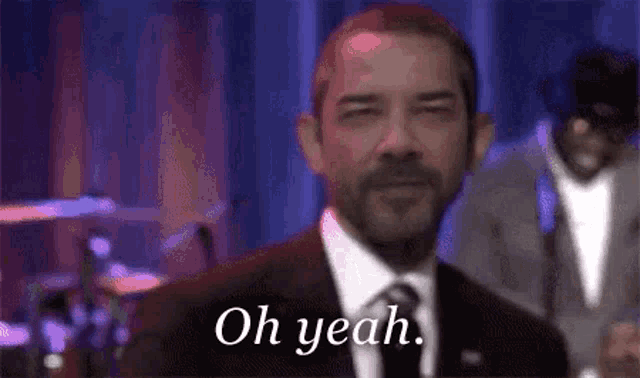 a man in a suit and tie is standing in front of a drum set and saying `` oh yeah '' .