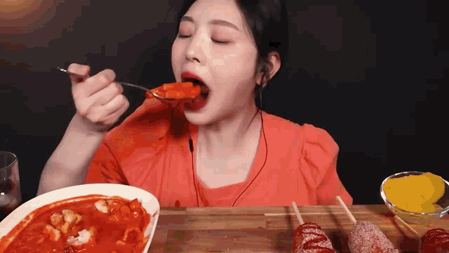a woman in an orange shirt is eating food with a spoon in her mouth .