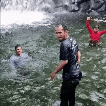 a man is standing in the water next to a waterfall while two other people are swimming in the water .
