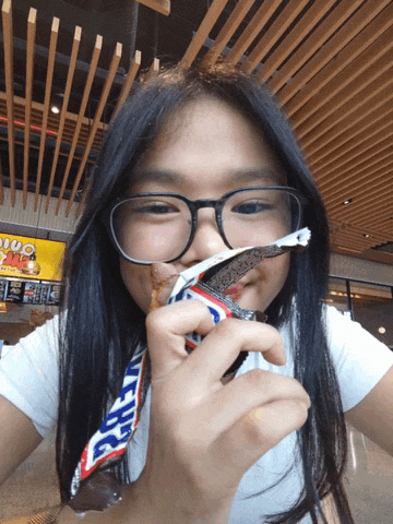 a girl with glasses is eating a snickers bar in a restaurant