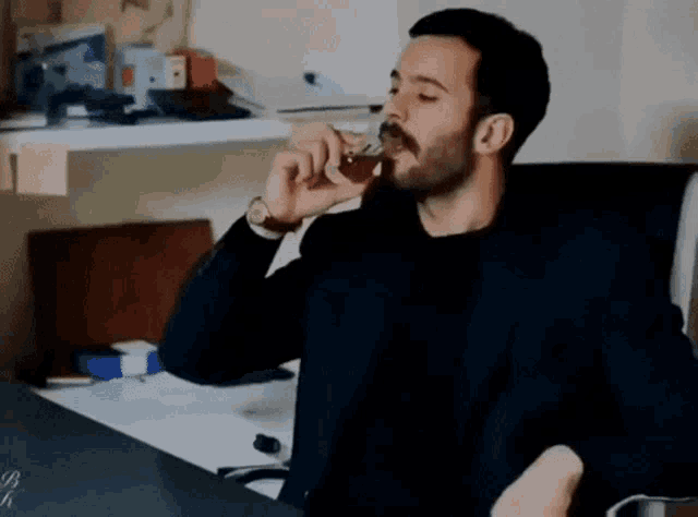 a man with a beard is sitting at a desk drinking from a glass