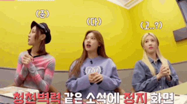 three girls are sitting at a table with their hands folded in front of a yellow wall with chinese writing on it .