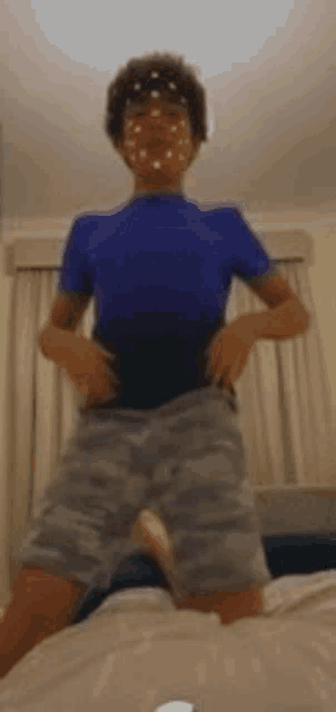 a boy in a blue shirt and camo shorts is standing on a bed .