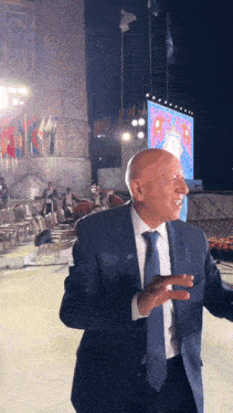 a bald man in a suit and tie stands in front of a large screen that says ua