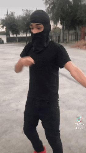 a man wearing a black t-shirt and a black balaclava is dancing in a parking lot .
