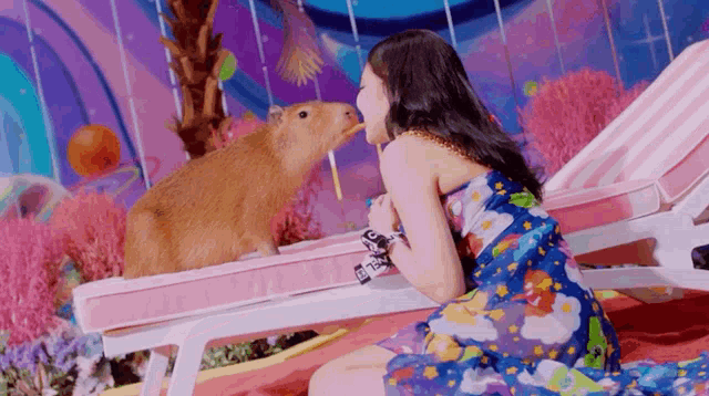 a woman in a blue dress is sitting next to a capybara eating a straw