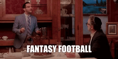 a man in a suit and tie is standing next to another man at a table and says fantasy football .