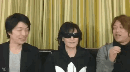 three men are sitting on a couch in front of a yellow curtain . one of the men is wearing sunglasses .