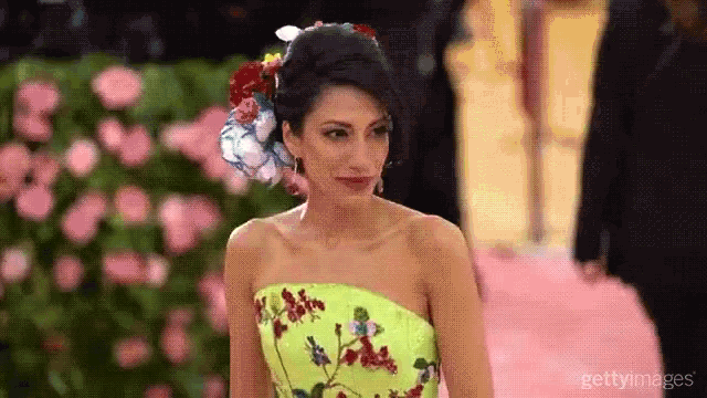 a woman in a yellow dress with flowers in her hair is standing in front of a wall of pink flowers .