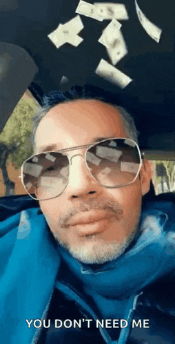 a man wearing sunglasses and a blue jacket is sitting in a car with money falling from the ceiling .