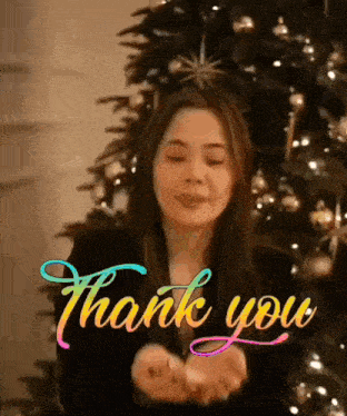 a woman is standing in front of a christmas tree and giving a thank you sign .