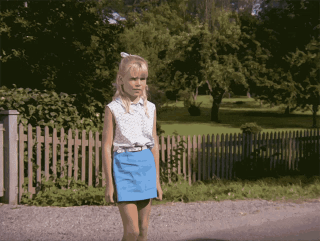 a girl wearing a blue skirt and a white shirt is standing in front of a wooden fence