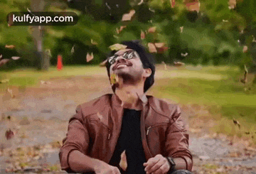 a man in a brown jacket is throwing leaves in the air in a park .