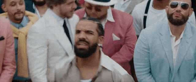 a man with a beard is sitting in a crowd of men wearing suits and sunglasses .