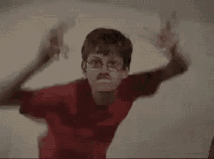 two young boys are dancing together in front of a white wall . one of the boys is wearing glasses and a red shirt .