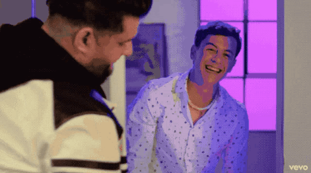 two men are laughing in front of a window and the word vevo is on the bottom left