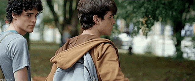 two young men standing next to each other in a park . one of the men is wearing a backpack .
