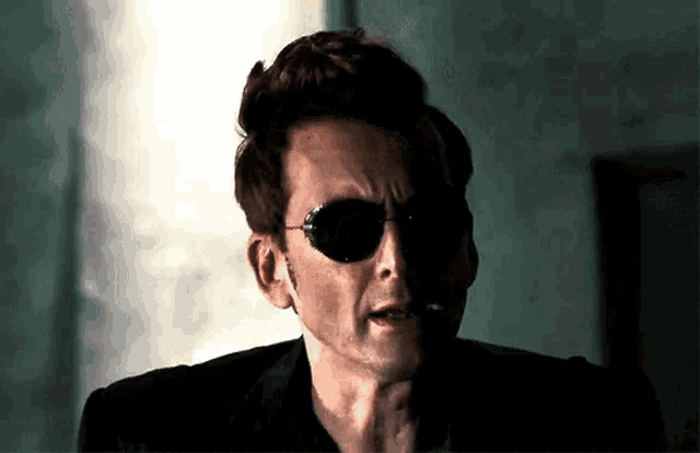 a man wearing sunglasses and a black suit is smoking a cigarette .
