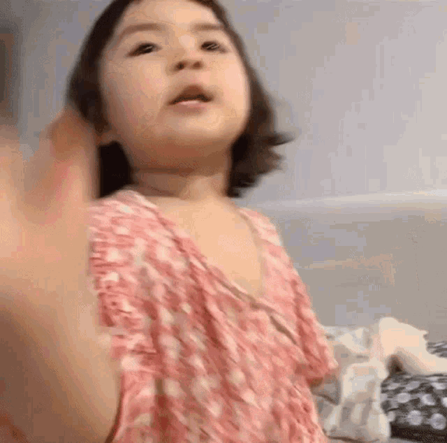a little girl in a pink dress is sitting on a bed and waving her hand .