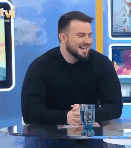 a man with a beard is sitting at a table with a glass of water in his hand .