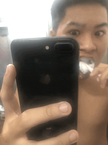 a shirtless man is brushing his teeth while taking a selfie with his iphone
