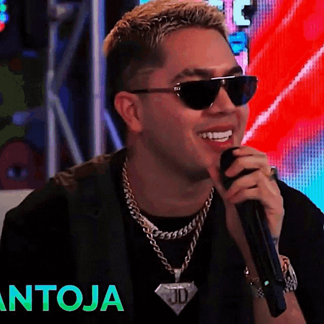 a man wearing sunglasses and a diamond pendant is singing into a microphone with the word antoja behind him
