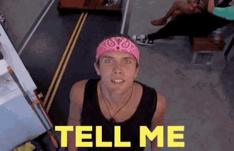 a man wearing a pink bandana is standing in front of a yellow sign that says tell me