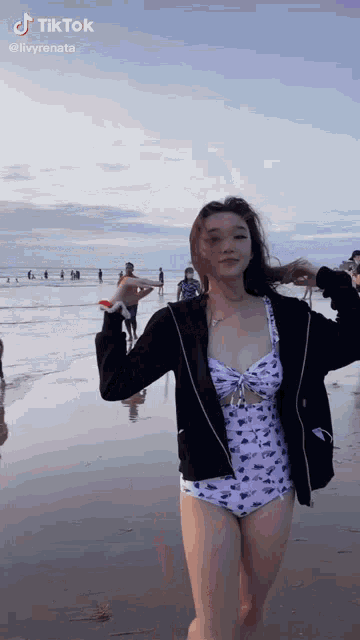 a woman in a swimsuit and a black jacket is standing on the beach .