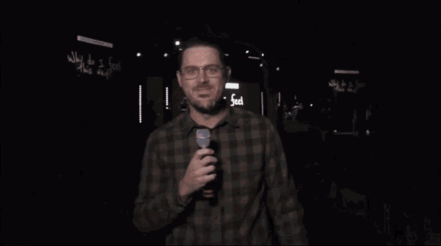 a man wearing glasses and a plaid shirt smiles in front of a sign that says why do i feel