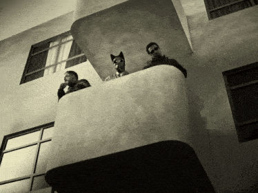 a man in a cat mask stands on a balcony with two other men