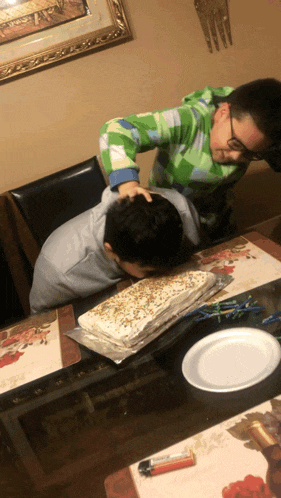 a boy in a green sweater holds another boy 's head over a cake on a table