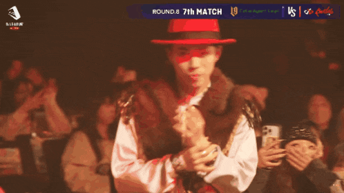 a man in a red hat stands in front of a crowd with the words round 8 7th match on the screen behind him